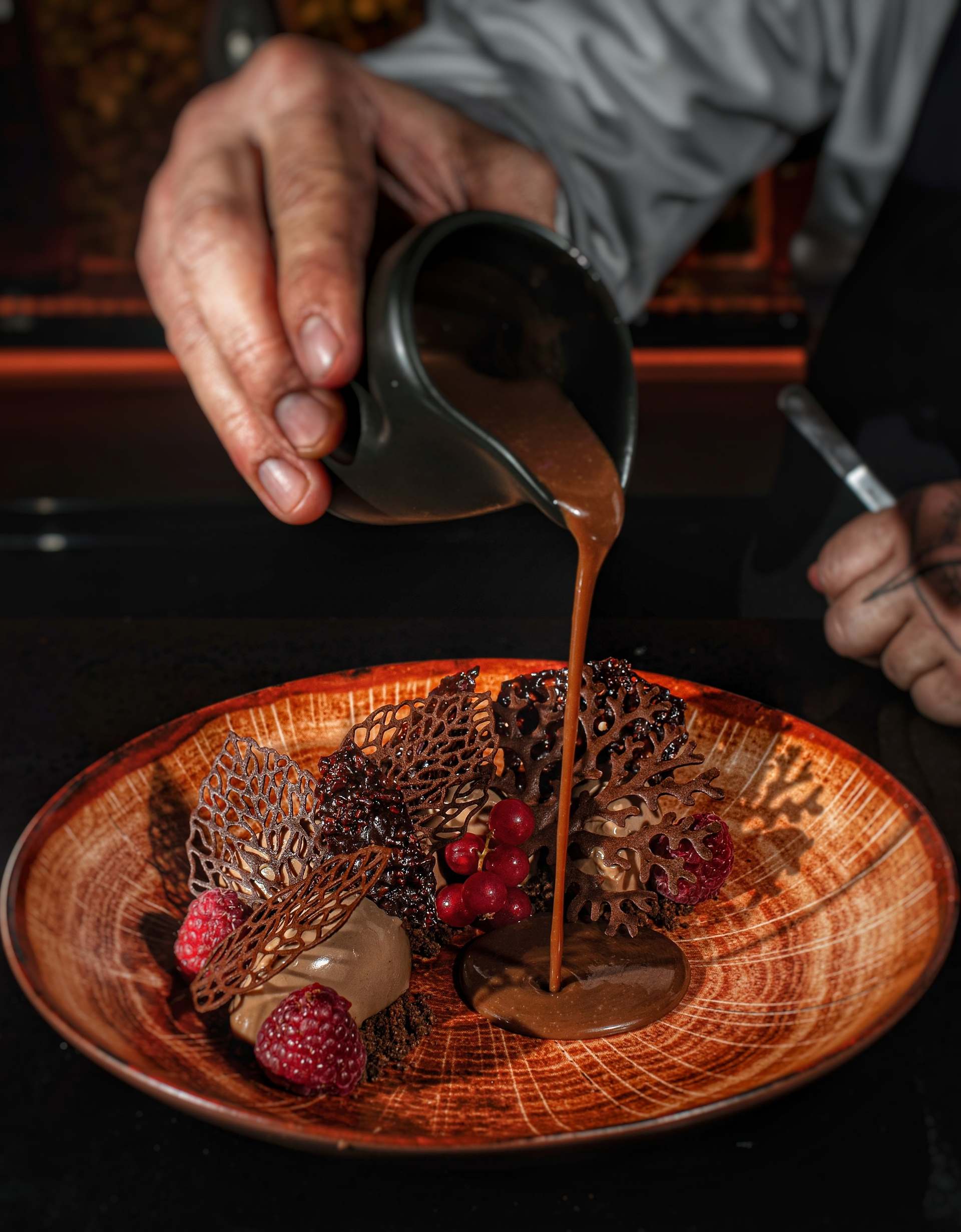 Black Forest dessert: a hand pours brown sauce onto an orange wood-grain plate beside lace tuiles, raspberries and redcurrants