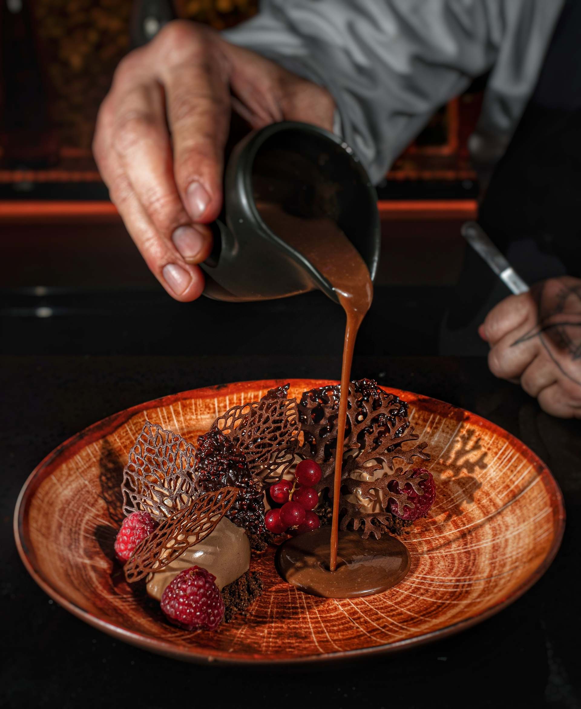Black Forest dessert: a hand pours brown sauce onto an orange wood-grain plate beside lace tuiles, raspberries and redcurrants