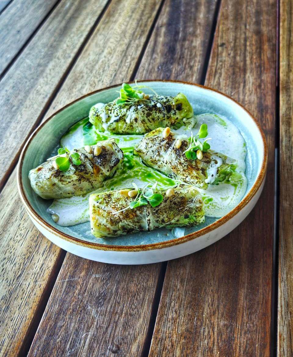 Four cabbage rolls with dark-speckled leaves on a white sauce with green oil, pine nuts and micro herbs in a grey-green bowl