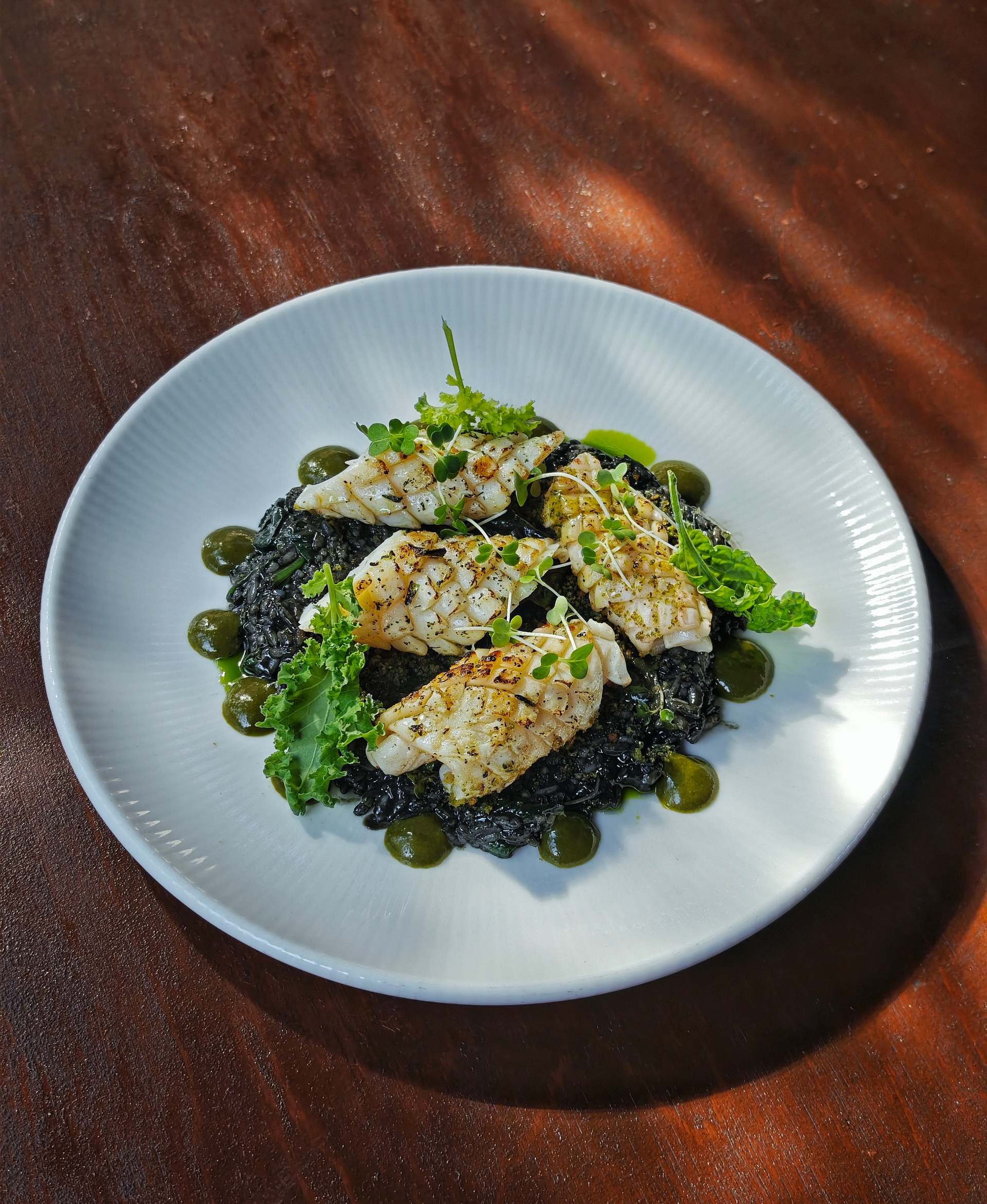 Four scored calamari pieces on black spinach rice with curly kale, micro herbs and dots of green purée on a white ribbed plate
