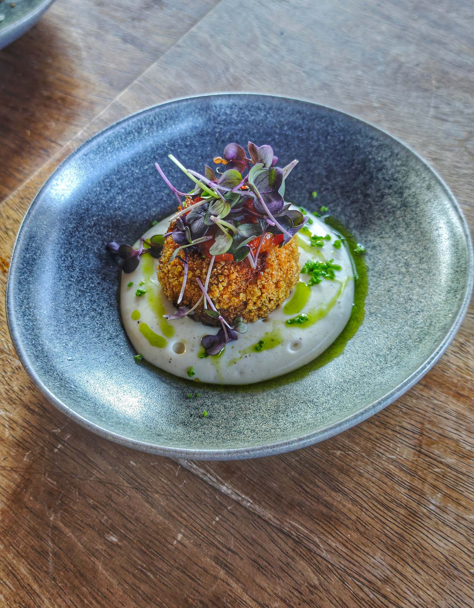 A round golden croqueta under purple micro herbs and a red topping, on white purée with green oil, in a speckled blue-grey bowl