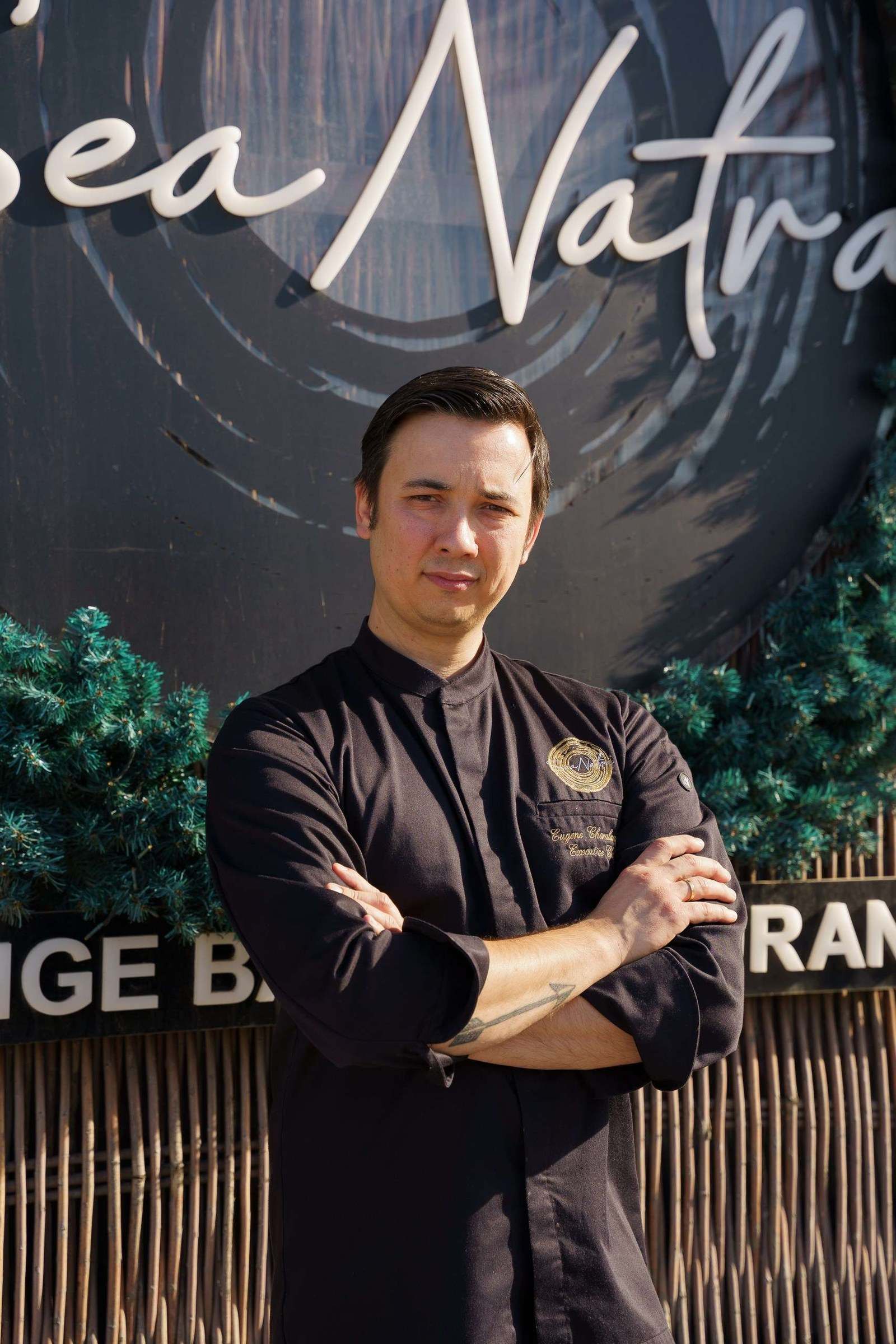 Ο Ευγένιος Χαραλάμπους, Executive Chef, με σκούρο σακάκι σεφ μπροστά σε μια επιγραφή εστιατορίου