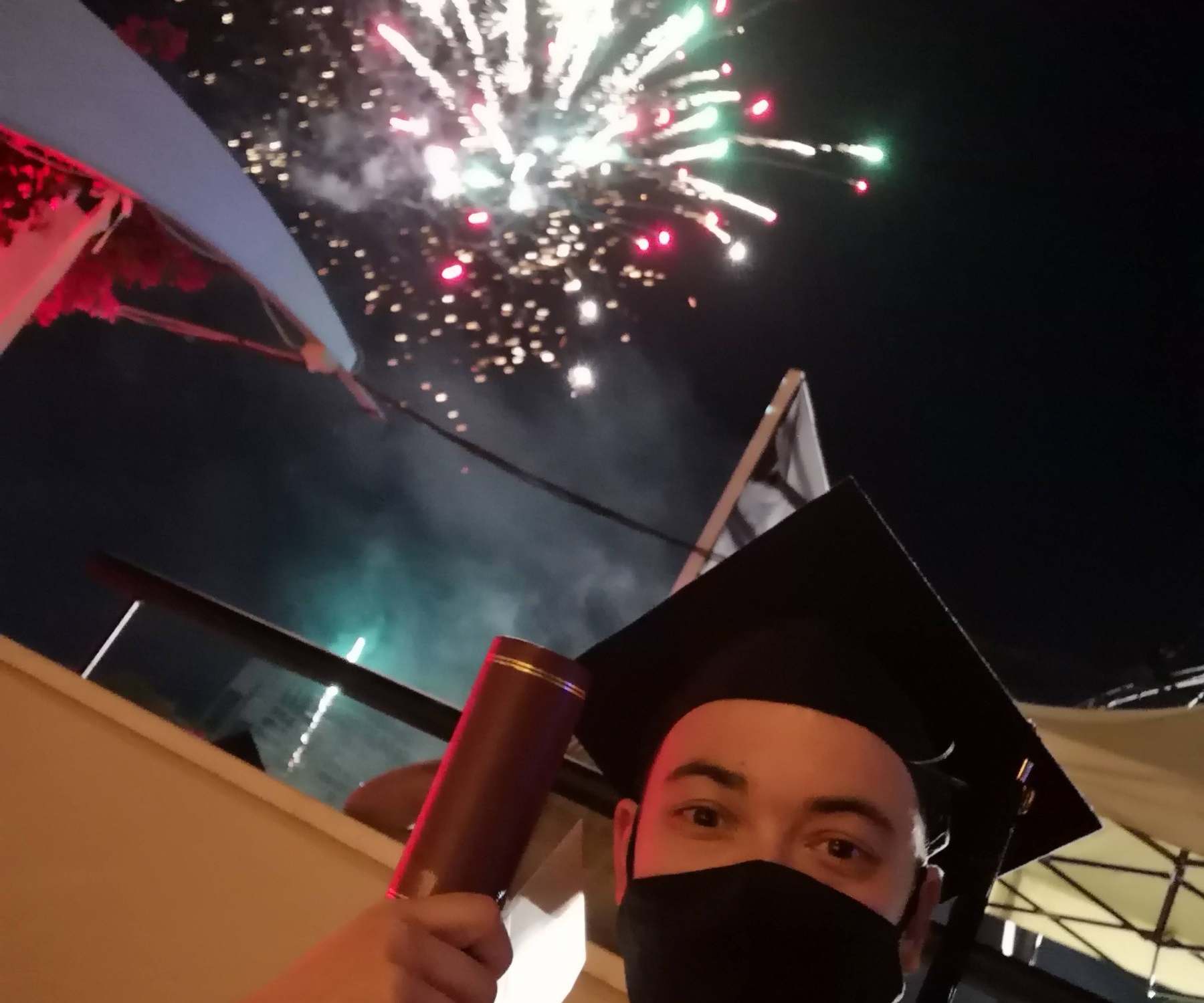 Eugene Charalampous in graduation cap and gown under fireworks at night