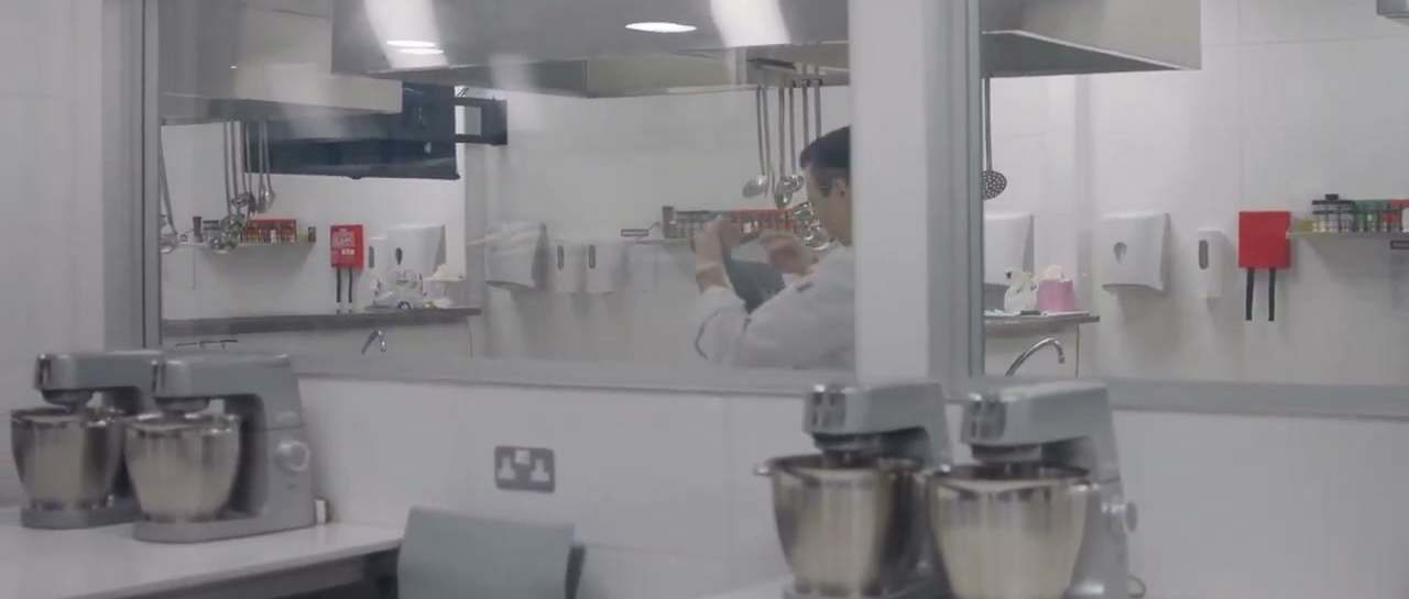 A spotless professional kitchen before service, stand mixers ready and a chef preparing behind the glass