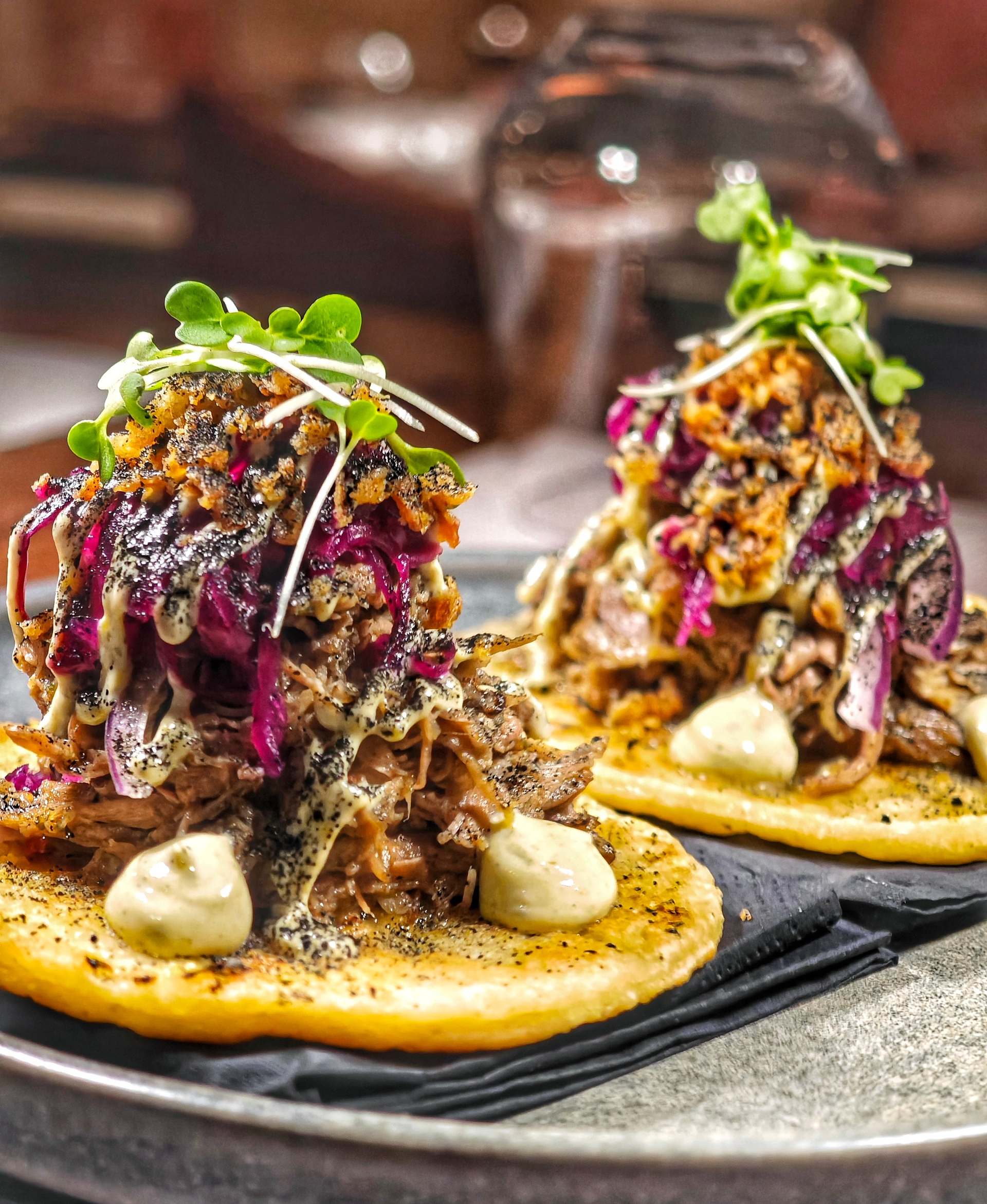 Two lamb gyros tacos on flat golden rounds piled with shredded meat, red cabbage, golden crumbs, pale sauce and micro herbs