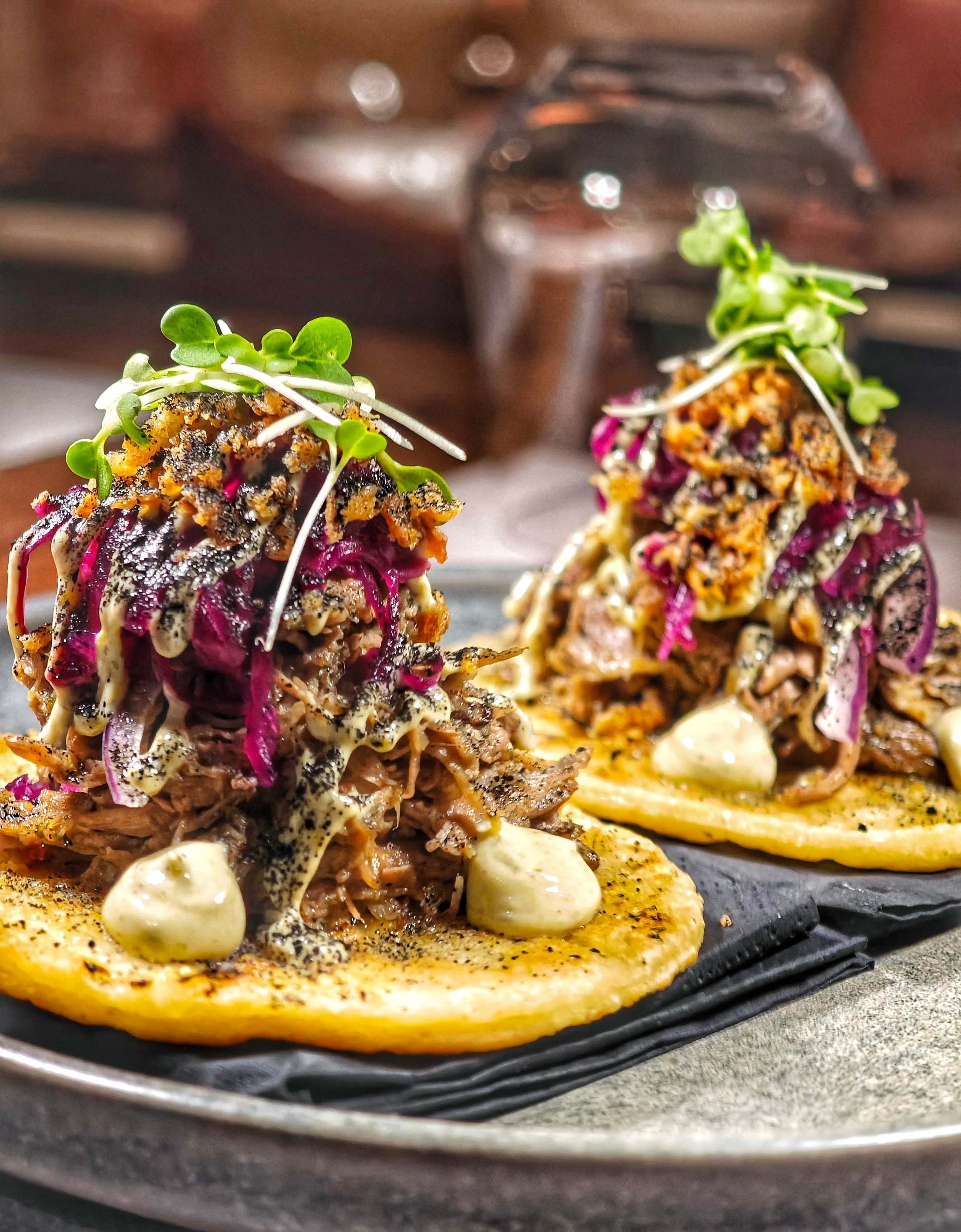 Two lamb gyros tacos on flat golden rounds piled with shredded meat, red cabbage, golden crumbs, pale sauce and micro herbs