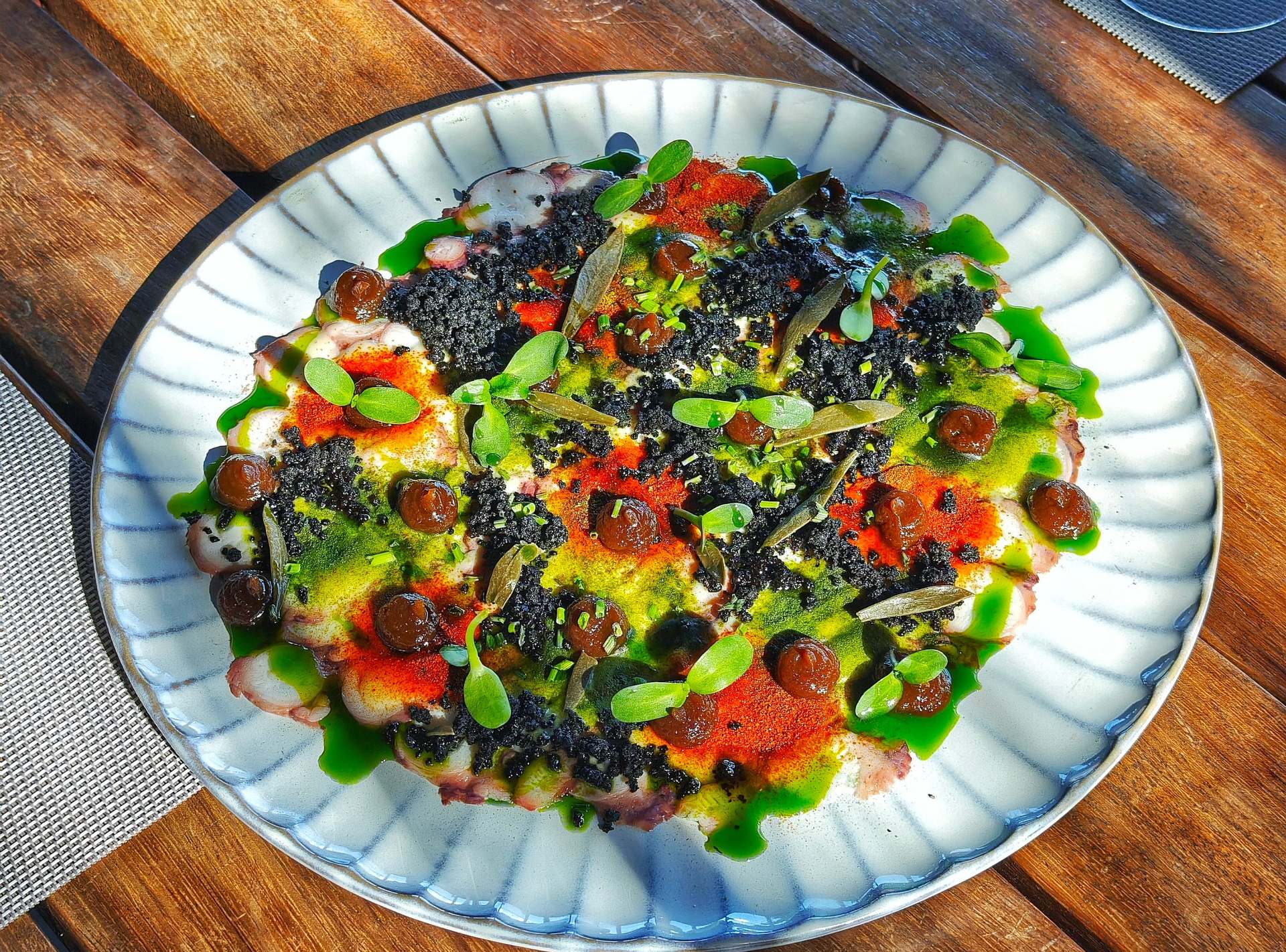 Карпаччо из осьминога под чёрной крошкой, красной и жёлто-зелёной пудрой, коричневыми точками, зелёным маслом и мелкими листьями, на рифлёной белой тарелке