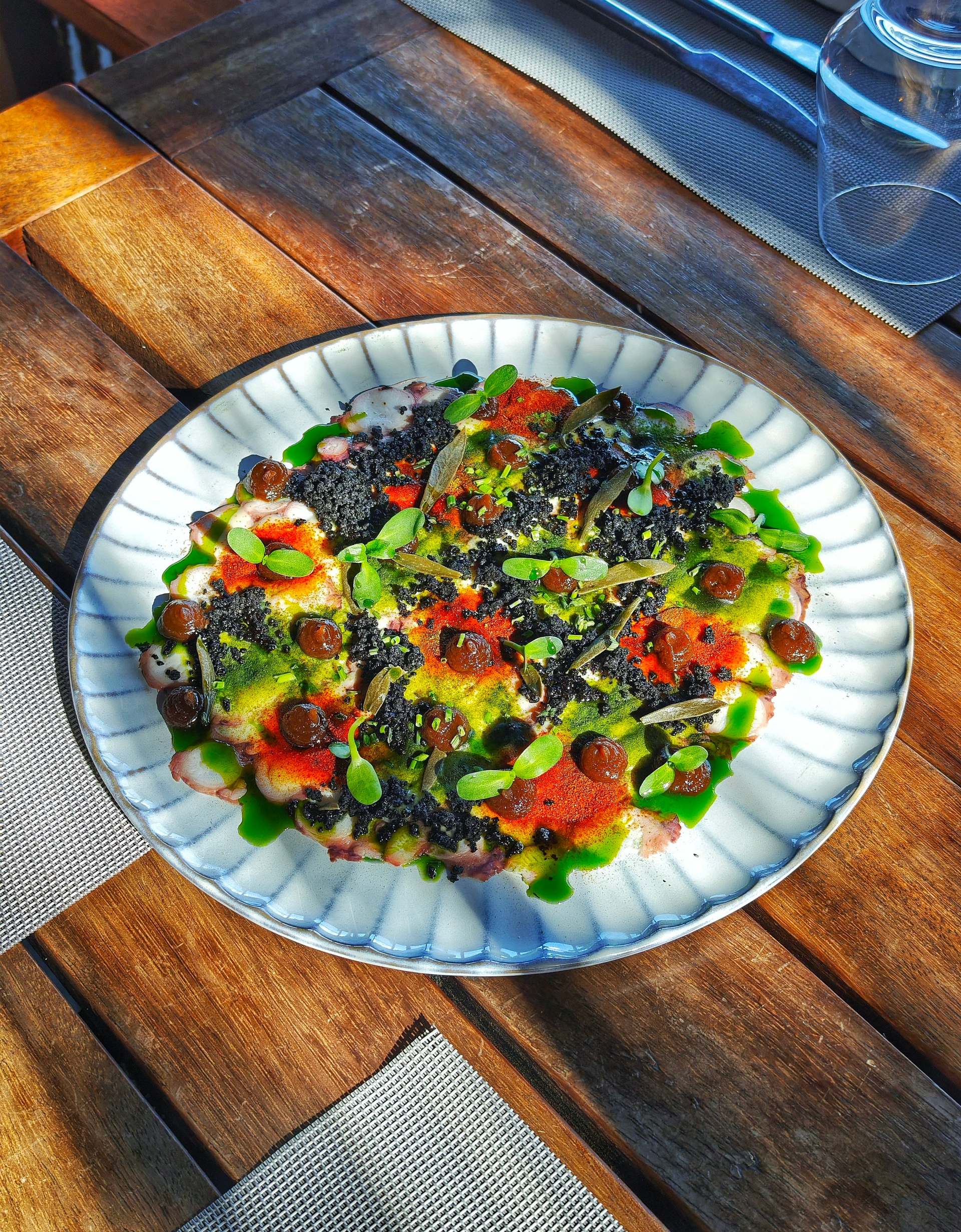 Octopus carpaccio under black crumb, red and yellow-green powders, brown dots, green oil and small leaves on a ribbed white plate