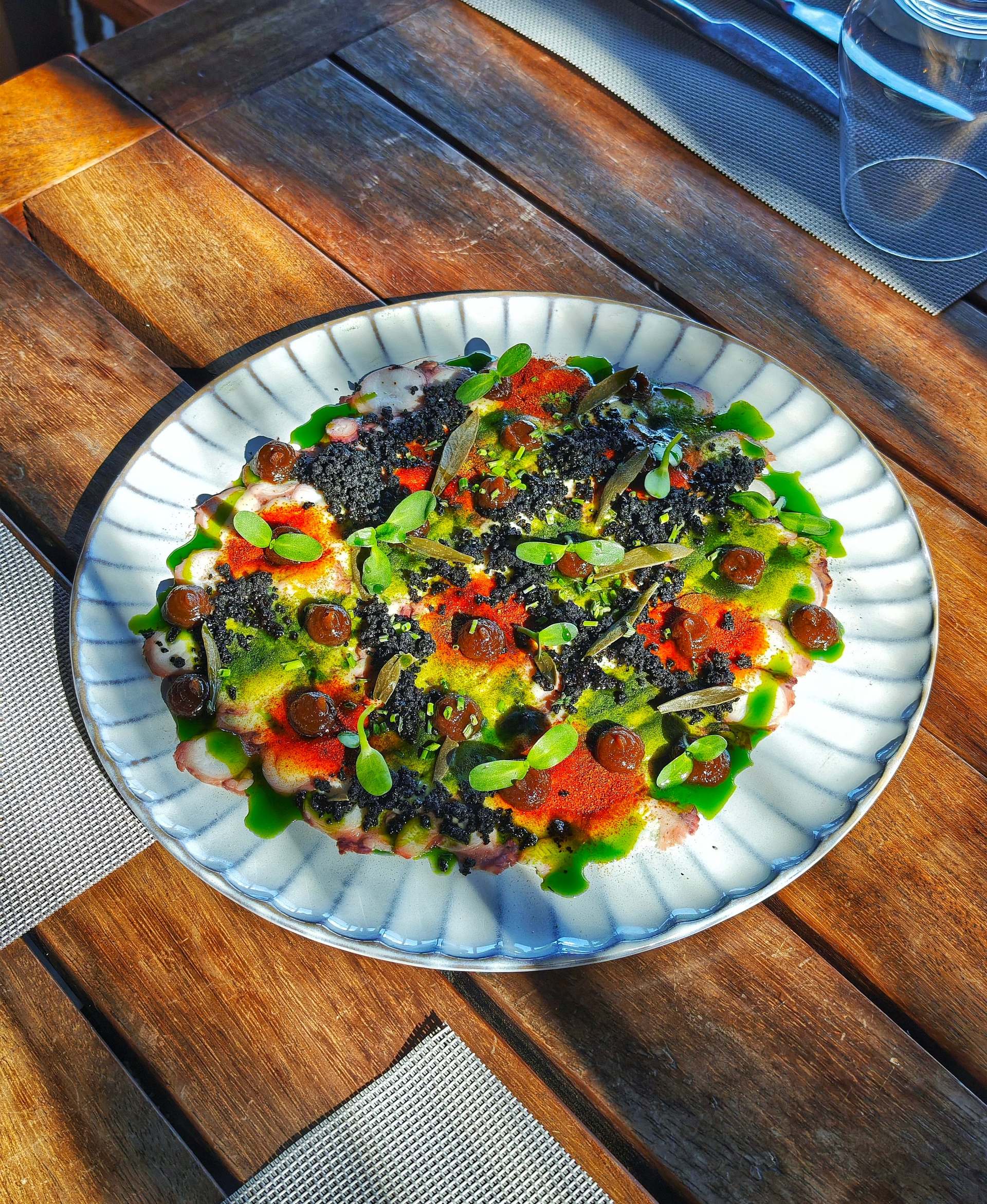 Octopus carpaccio under black crumb, red and yellow-green powders, brown dots, green oil and small leaves on a ribbed white plate