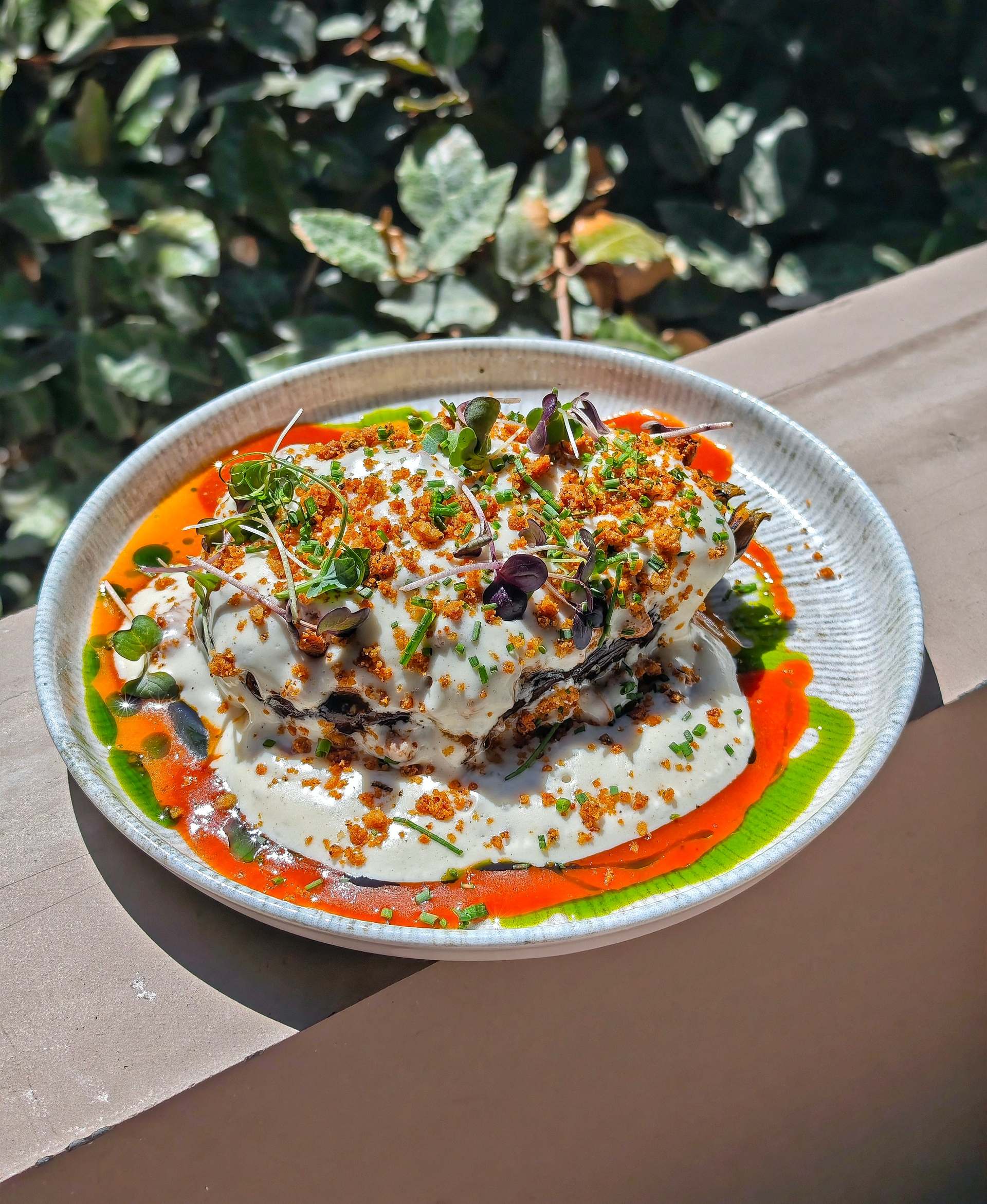 Papoutsaki aubergine under thick white sauce, orange crumb, chives and micro herbs, with red sauce and green oil on a pale plate