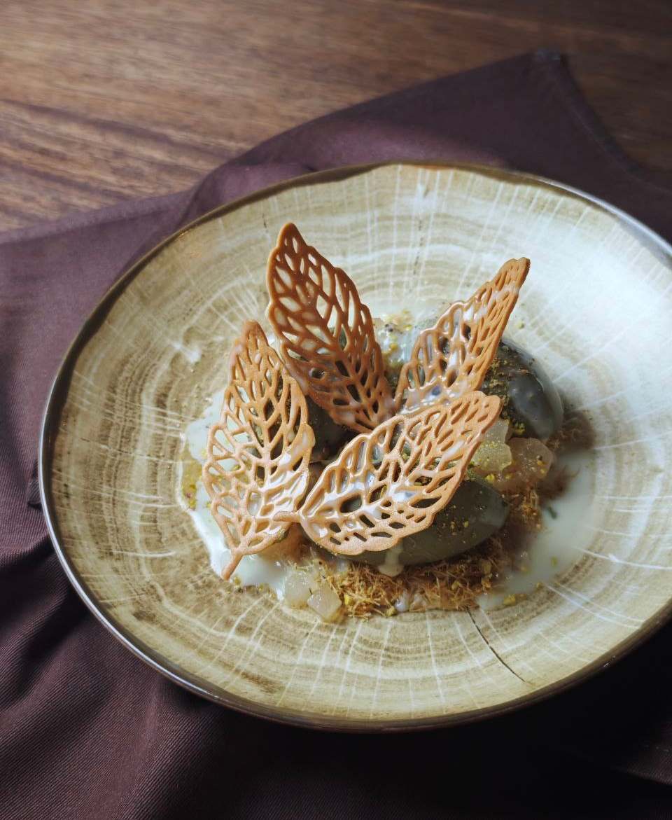 Pistachio Bliss: four lace leaves over grey-green quenelles, white sauce and golden strands in a bowl with a wood-grain glaze