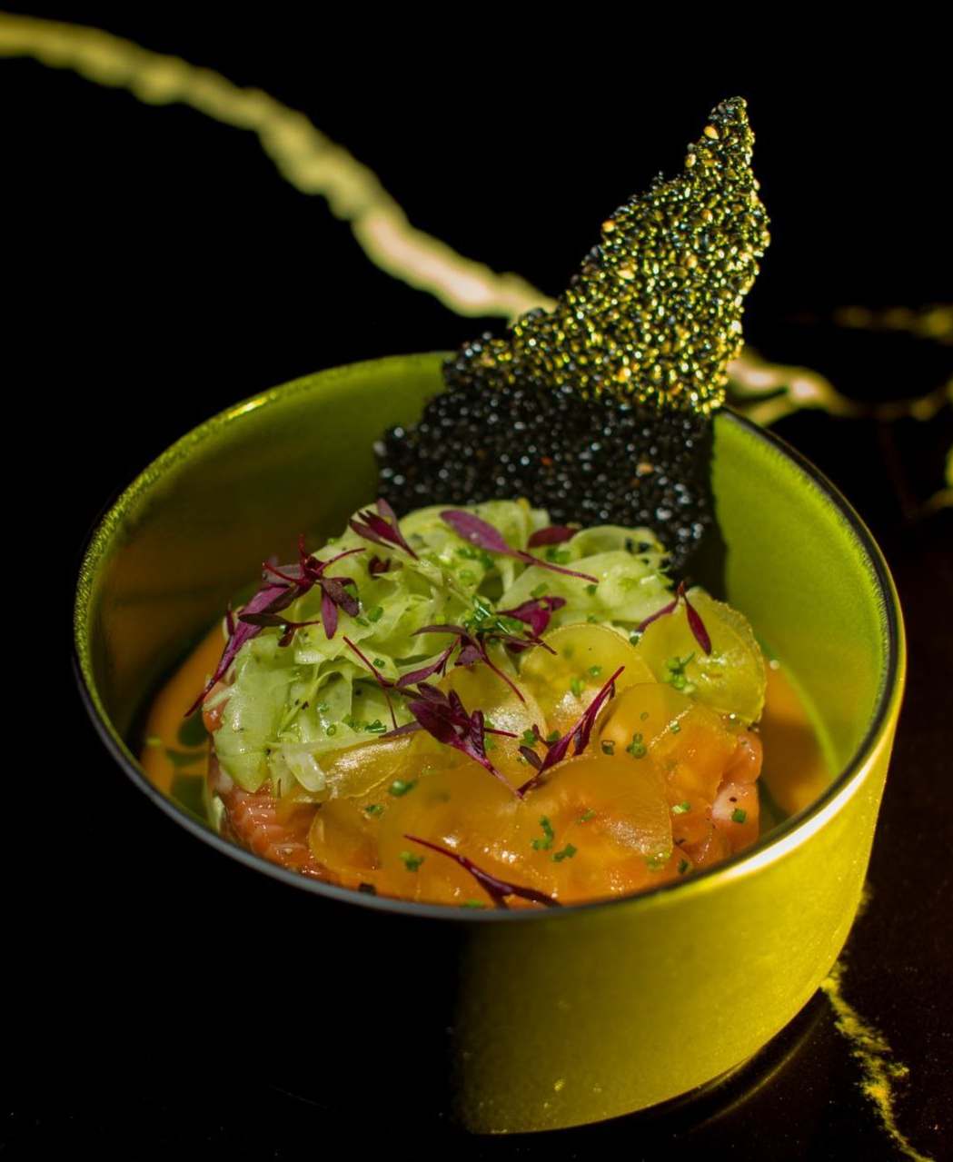 Salmon tartare under fennel ribbons, thin golden slices and purple micro herbs, with a black seeded crisp behind, in a green bowl
