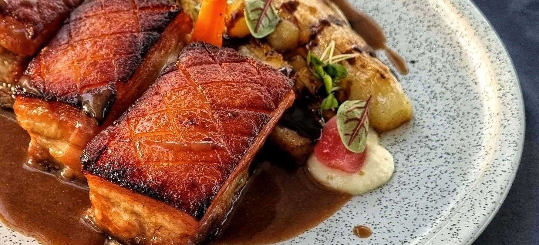 Crisp glazed pieces of smoked pork belly with jus, vegetables and parsnip cream on a speckled plate
